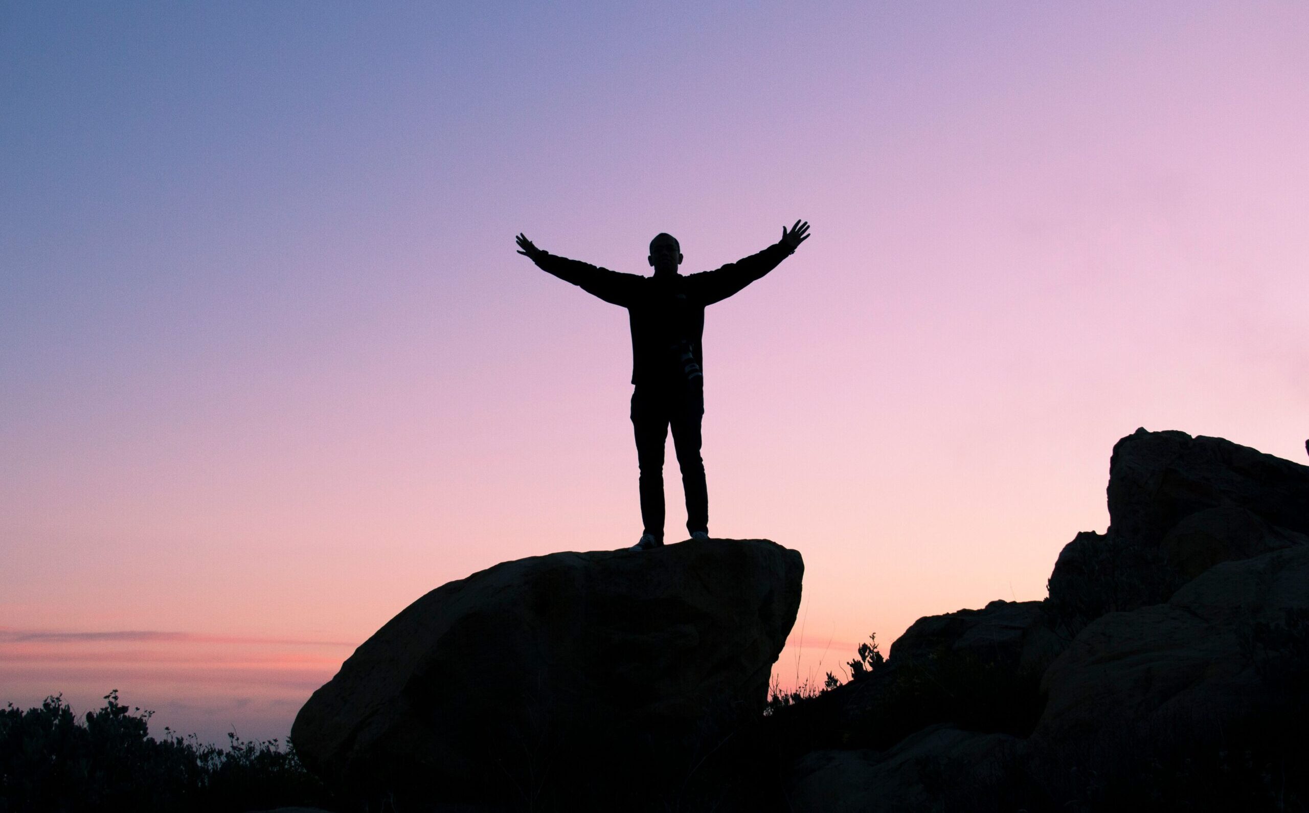 man standing on top of a hill with his arms up at sunset.